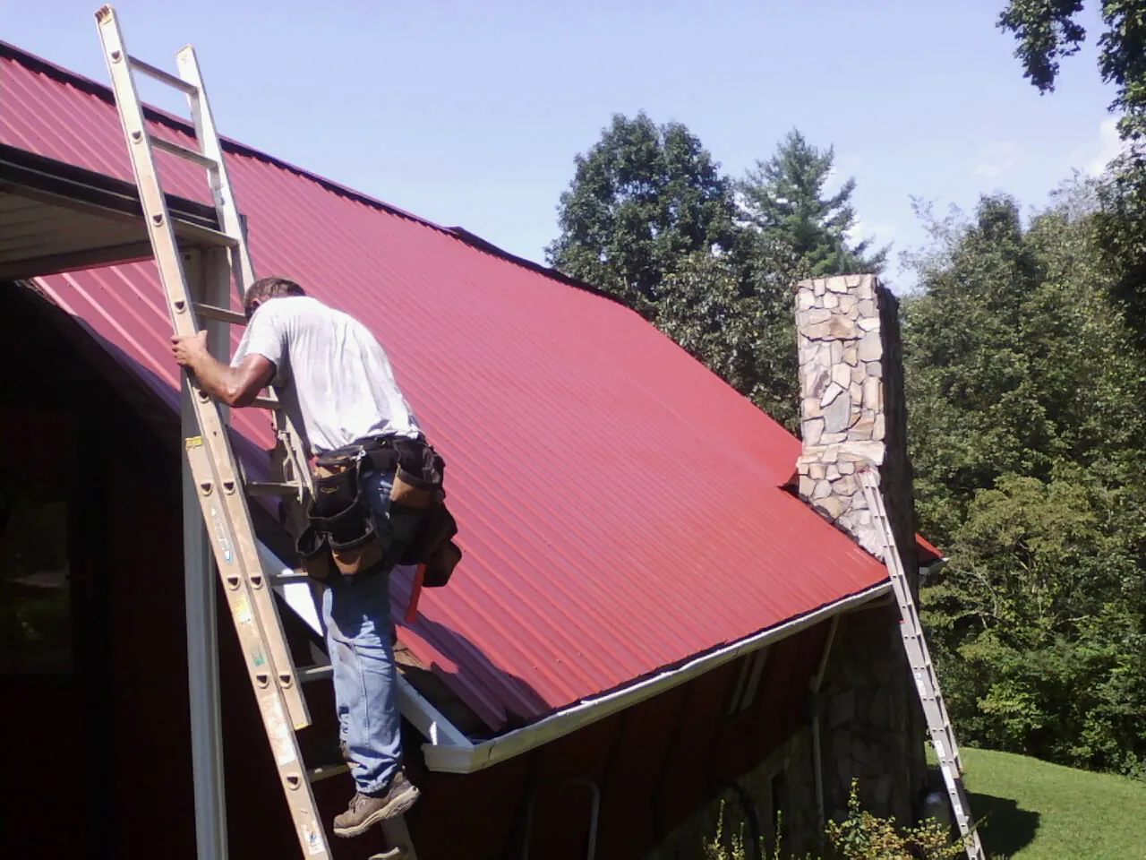 Roofer on a ladder beside a red metal roof and stone chimney — Soffit & Fascia Repair by Roofing Brothers in Marblemount